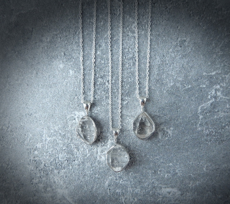 May include: Four silver necklaces with clear crystal pendants hanging against a grey textured background.
