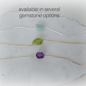 May include: Three delicate necklaces with different colored gemstones. The necklaces are on a white marble surface. The text "available in several gemstone options" is above the necklaces.