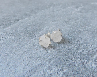 Puede incluir: Un par de pendientes de botón pequeños y redondos. Cada pendiente presenta una piedra de color claro, de corte rugoso, engastada en una base de metal plateado. Los pendientes se muestran sobre una superficie gris texturizada.