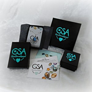 May include: A black box with the text "GSA GemStoneAppeal" in turquoise lettering and a turquoise heart. The box is sitting on a white surface. There are two other boxes with the same logo and a small card with the same logo and the text "Care Tip: A little love goes a long way..." in turquoise lettering.