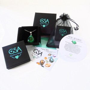 May include: A green malachite gemstone pendant necklace in a black box with a white background. The necklace is packaged with a black velvet bag and a card with information about the stone. The card reads "Malachite, prosperity, protection, renewal, abundance".