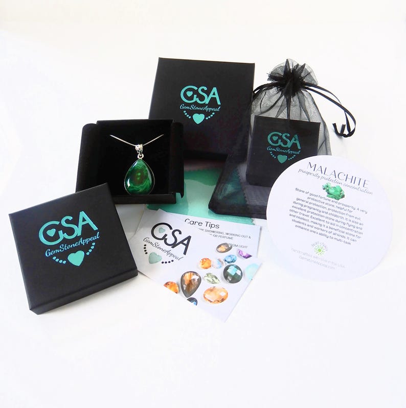 May include: A green malachite gemstone pendant necklace in a black box with a white background. The necklace is packaged with a black velvet bag and a card with information about the stone. The card reads "Malachite, prosperity, protection, renewal, abundance".
