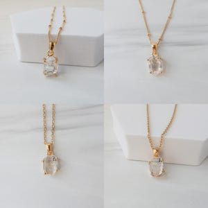 May include: Four gold necklaces, each featuring a clear, oval pendant. The pendants are set in gold and hang from delicate gold chains. The necklaces are displayed on white geometric blocks. The gemstones appear to be faceted. The chains have small gold beads.