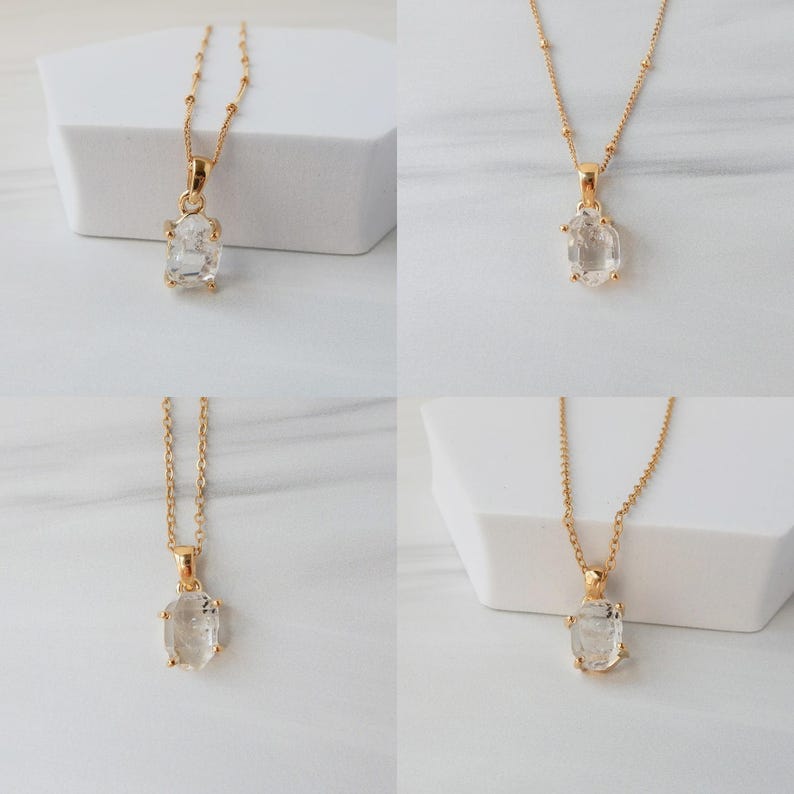 May include: Four gold necklaces, each featuring a clear, oval pendant. The pendants are set in gold and hang from delicate gold chains. The necklaces are displayed on white geometric blocks. The gemstones appear to be faceted. The chains have small gold beads.