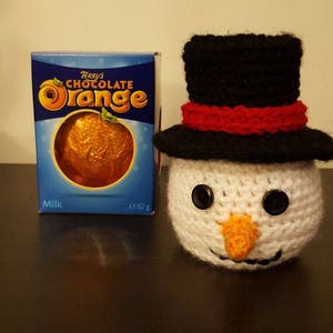 May include: A handmade crocheted snowman with a black top hat and a red band, black button eyes, and an orange carrot nose. Next to it is a box of Terry's Chocolate Orange, a milk chocolate treat, weighing 157g.