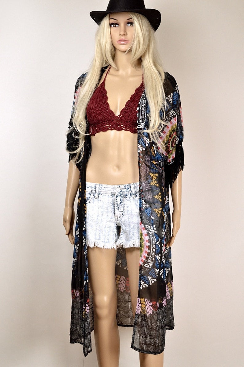 May include: A black sheer kimono with a colorful floral pattern and fringe trim. The kimono is worn over a red crochet top and denim shorts.