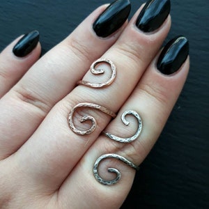 Puede incluir: Tres anillos de espiral plateados y dorados en una mano con esmalte de uñas negro. Los anillos son ajustables y se pueden usar en cualquier dedo.