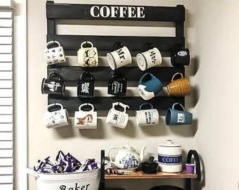 May include: A black wooden coffee mug rack with the word "COFFEE" in white letters. The rack is mounted on a wall and has 12 hooks for hanging mugs. There are various mugs hanging on the hooks, including some with designs and text. Below the rack is a wooden shelf with a coffee maker, a coffee container, a teapot, and a white metal container with the word "Baker" on it.