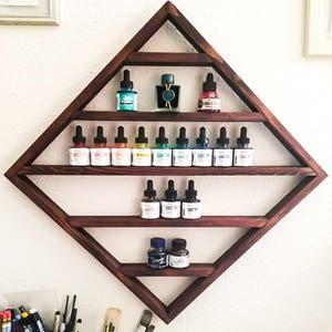 May include: A dark wood diamond-shaped shelf with multiple shelves holding small bottles of colorful ink. The shelf is mounted on a white wall.