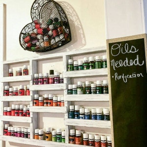 May include: A white wooden shelf unit with multiple rows of small glass bottles filled with colorful liquids. The bottles are arranged in a grid pattern. A heart-shaped wire basket filled with more bottles is hanging above the shelf. A chalkboard sign to the right reads "Oils Needed - Purification."