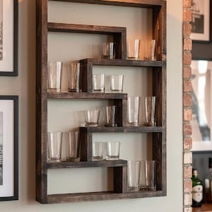 May include: A rectangular dark brown wooden wall shelf with an asymmetrical, multi-level interior design. The shelf features several horizontal surfaces holding a collection of clear glass tumblers of varying heights, arranged in a staggered pattern.