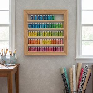 May include: A wooden framed display case filled with colorful paint tubes, arranged in a rainbow spectrum. The case is mounted on a light gray wall. A wooden table with art supplies and a wire basket with rolled paper are in the foreground.
