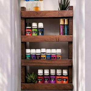May include: A wooden wall-mounted shelf with four tiers, holding various essential oil bottles. The bottles are arranged in rows and are labeled with different names, such as "Tranquility", "Deep Relief", "Stress Away", and "Cypress". There are also two small potted succulents on the shelf.