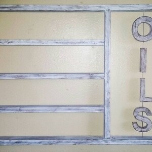 Puede incluir: Una estanter&iacute;a de pared de madera blanca envejecida con tres estantes horizontales y una secci&oacute;n vertical. La palabra "OILS" cuelga verticalmente en la secci&oacute;n derecha. Los estantes est&aacute;n pintados de blanco con tonos grises.