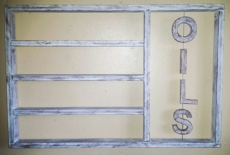 Puede incluir: Una estanter&iacute;a de pared de madera blanca envejecida con tres estantes horizontales y una secci&oacute;n vertical. La palabra "OILS" cuelga verticalmente en la secci&oacute;n derecha. Los estantes est&aacute;n pintados de blanco con tonos grises.