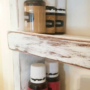 Op de afbeelding: Een wit houten plank met drie rijen kleine bruine glazen flessen met witte deksels. De flessen hebben etiketten met de woorden "Copaiba" en "Ylang Ylang".