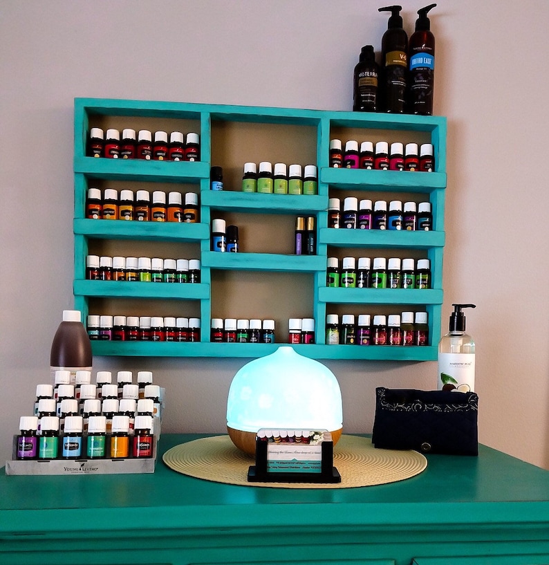Op de afbeelding: Een turquoise houten rek met meerdere rijen kleine glazen flesjes gevuld met etherische oli&euml;n. Het rek is aan een muur bevestigd en heeft een diffuser en andere flesjes op een turquoise tafel eronder.