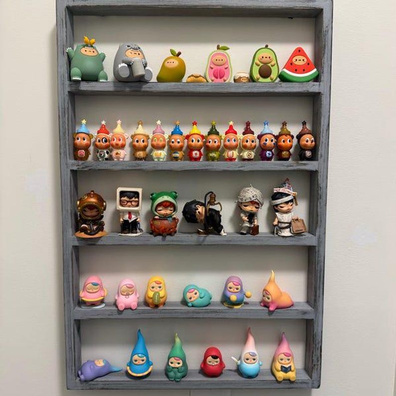 May include: A rectangular grey wooden display shelf with five horizontal tiers holding a collection of small, colourful vinyl character figurines. The figures vary in shape, including fruit-themed, animal-inspired, and human-like designs.