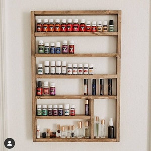 May include: A wooden essential oil shelf filled with various essential oil bottles. The shelf is made of light brown wood and is mounted on a white wall. The bottles are of different sizes and colors, with white and colored labels.