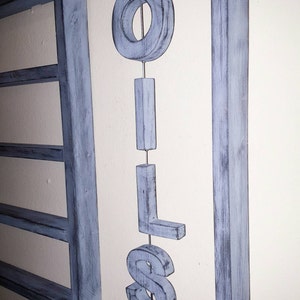 Puede incluir: Un letrero de madera azul claro con la palabra "OILS" cuelga de un alambre de metal. Las letras son tridimensionales y est&aacute;n suspendidas frente a un marco de madera azul a juego.