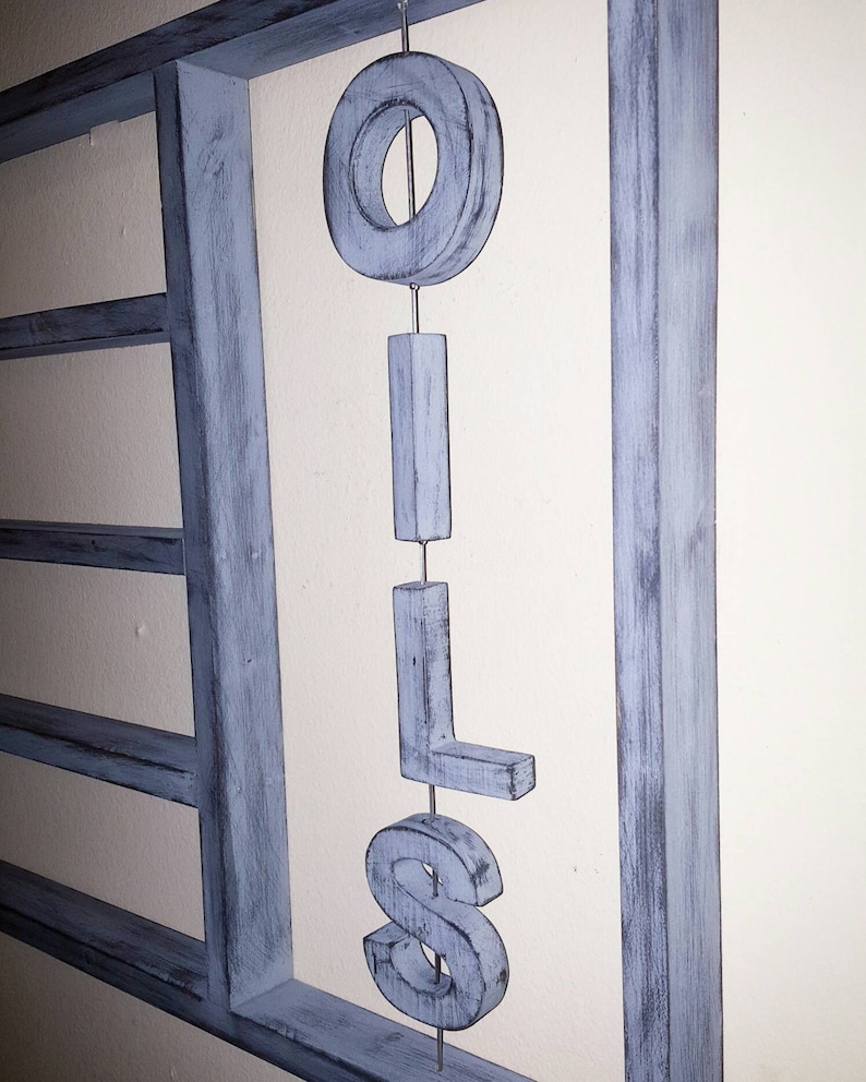 Puede incluir: Un letrero de madera azul claro con la palabra "OILS" cuelga de un alambre de metal. Las letras son tridimensionales y est&aacute;n suspendidas frente a un marco de madera azul a juego.