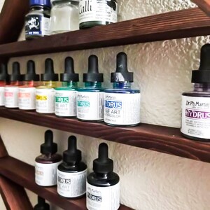 May include: A wooden shelf unit with multiple shelves displaying bottles of Dr. Ph Martin's Hydrus Fine Art Watercolors in various colors. The bottles have dropper tops and are arranged in rows on the shelves.
