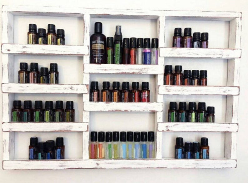 May include: White wooden shelf unit with multiple shelves, each filled with small glass bottles of essential oils. The bottles are arranged in rows and are labeled with different names. The shelf unit is a great way to store and display essential oils.