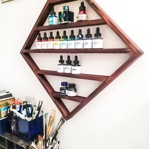 May include: A dark wood diamond-shaped shelf with five shelves holding small bottles of colorful liquid. The shelf is mounted on a white wall.