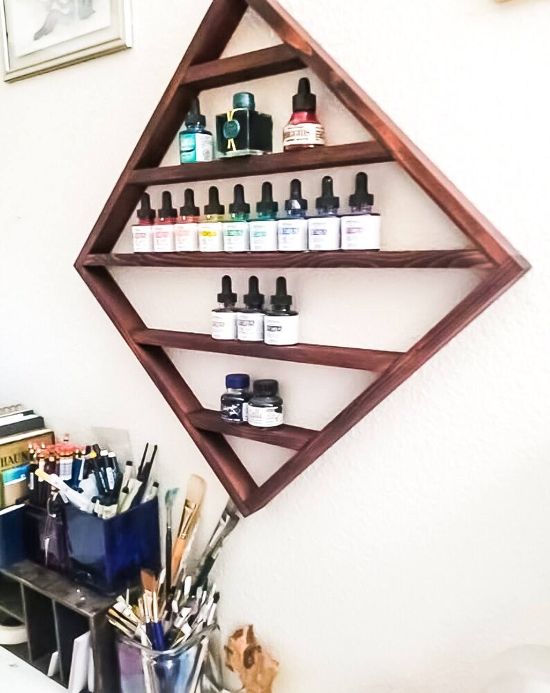 May include: A dark wood diamond-shaped shelf with five shelves holding small bottles of colorful liquid. The shelf is mounted on a white wall.