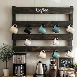 May include: A dark brown wooden wall-mounted mug rack with the word Coffee printed in white. The rack features multiple black metal hooks holding various ceramic mugs in shades of cream, black, and blue. Below the rack sits a coffee station.