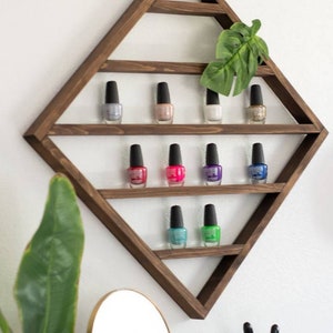 May include: A wooden diamond-shaped shelf with multiple shelves to display nail polish bottles. The shelf is mounted on a white wall. There are 9 bottles of nail polish on the shelves, in various colours, including pink, blue, green, and purple.