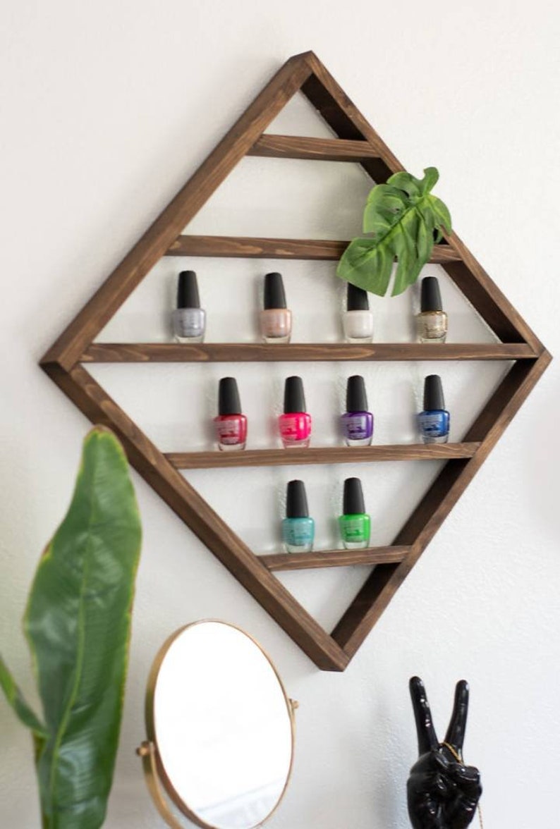 May include: A wooden diamond-shaped shelf with multiple shelves to display nail polish bottles. The shelf is mounted on a white wall. There are 9 bottles of nail polish on the shelves, in various colours, including pink, blue, green, and purple.