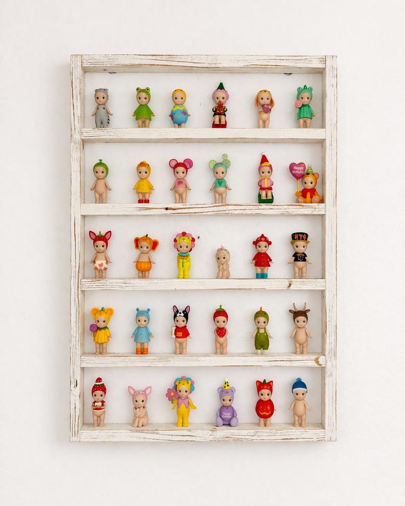 May include: A white wooden shelf displaying a collection of small, colorful figurines. The figurines are dressed in various costumes, including animals, fruits, and other whimsical designs. The shelf has multiple levels, each holding several figurines.