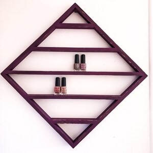 May include: A purple diamond-shaped shelf with five shelves for displaying nail polish bottles. The shelf is mounted on a white wall. Three bottles of nail polish are displayed on the top shelf and two bottles are displayed on the second shelf.