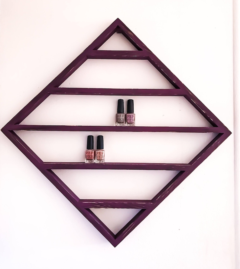 May include: A purple diamond-shaped shelf with five shelves for displaying nail polish bottles. The shelf is mounted on a white wall. Three bottles of nail polish are displayed on the top shelf and two bottles are displayed on the second shelf.