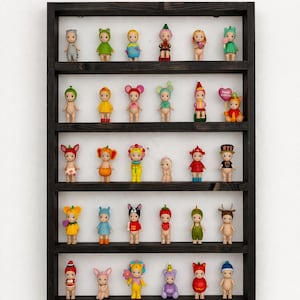 May include: A dark wooden display shelf filled with small, colorful figurines. The figurines are dressed in various outfits, including animal costumes and floral designs. The shelf is mounted on a white wall, creating a clean and organized presentation of the collectibles.