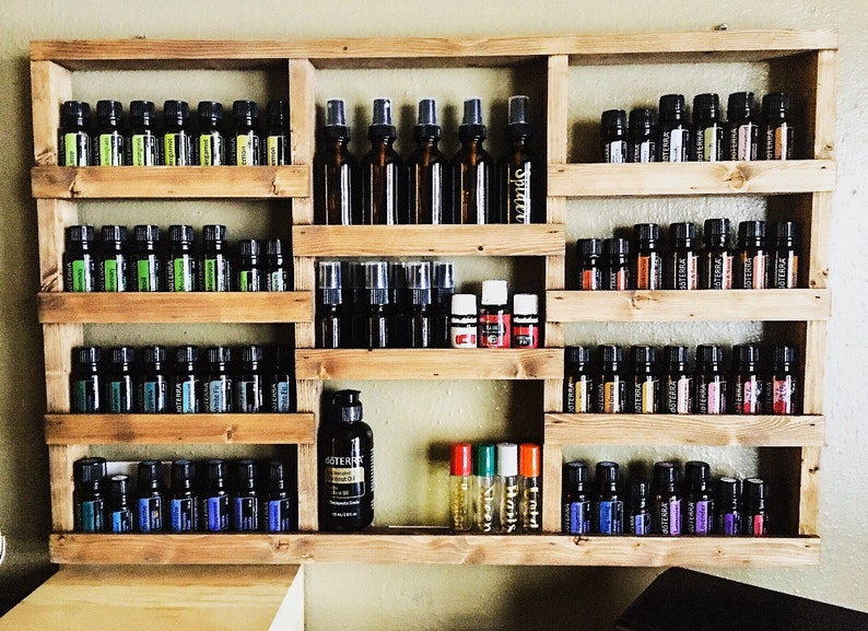 May include: A wooden shelf unit with multiple shelves, each filled with small glass bottles of essential oils. The bottles are arranged in rows and are of various colors, including green, blue, and purple. There are also a few larger bottles with spray tops.