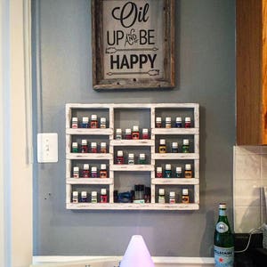 Op de afbeelding: Een wit houten plank met meerdere kleine glazen flesjes gevuld met kleurrijke vloeistoffen. De plank is gemonteerd aan een grijze muur met een ingelijst bord waarop staat "Oil Up and Be Happy". Een diffuser met een paars licht staat op een aanrecht onder de plank.