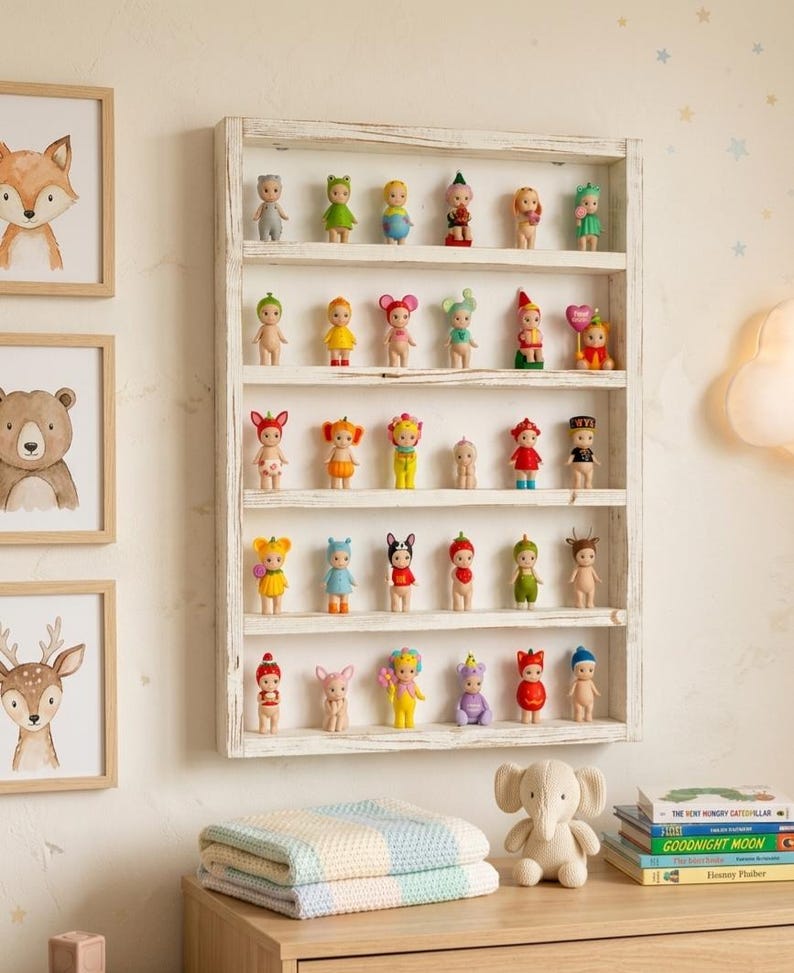 May include: A white, distressed wooden shelf displays a collection of small, colorful figurines. The figurines are anthropomorphic, with various outfits and accessories. The shelf is mounted on a wall with framed animal art and a stack of books.