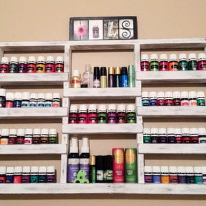 May include: White wooden shelf unit with multiple shelves holding rows of small glass bottles with white lids. The bottles are filled with colorful liquids. The shelf unit is mounted on a wall.