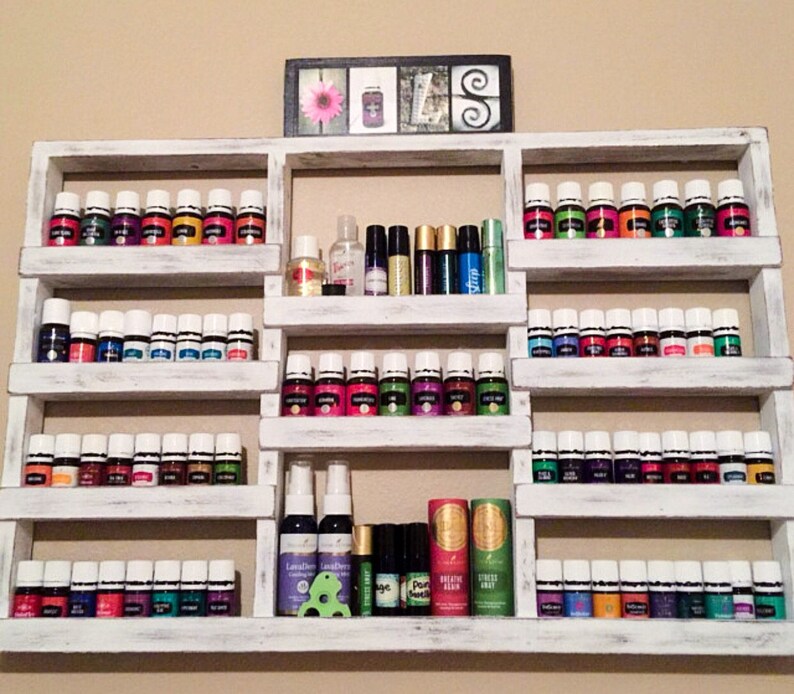 May include: White wooden shelf unit with multiple shelves holding rows of small glass bottles with white lids. The bottles are filled with colorful liquids. The shelf unit is mounted on a wall.
