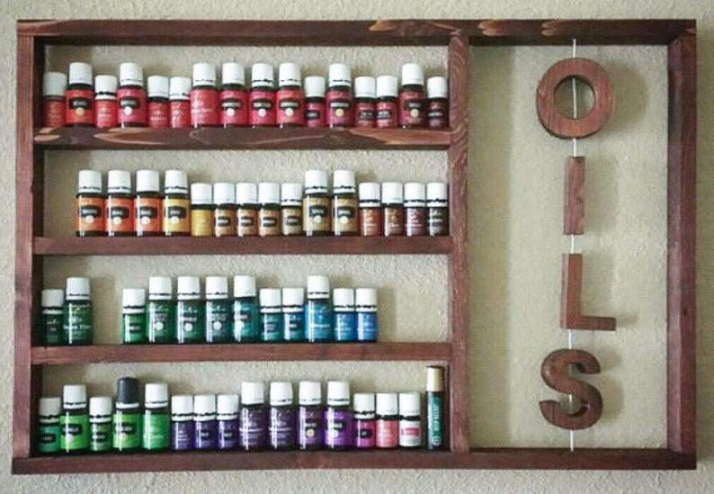 Puede incluir: Estante de madera para aceites esenciales con m&uacute;ltiples filas de peque&ntilde;os frascos de vidrio con tapas blancas. Los frascos est&aacute;n llenos de aceites de varios colores. La palabra "OILS" est&aacute; escrita con letras de madera colgando a la derecha.