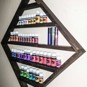 May include: A dark brown wooden triangular shelf with three shelves, holding rows of small glass bottles with white caps. The bottles are filled with colourful liquids. The shelf is mounted on a white wall.