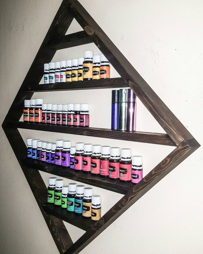 May include: A dark brown wooden triangular shelf with three shelves, holding rows of small glass bottles with white caps. The bottles are filled with colourful liquids. The shelf is mounted on a white wall.