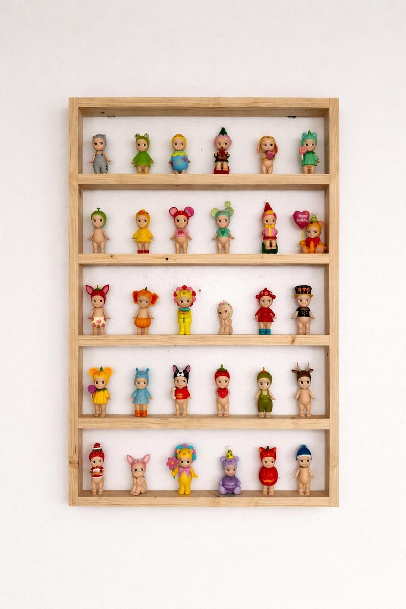 May include: A wooden display shelf filled with small, colorful figurines. The figurines are dressed in various outfits, including fruits, animals, and other whimsical designs. The shelf is made of light-colored wood and is mounted on a white wall.