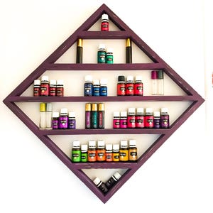 May include: A purple diamond-shaped shelf with five shelves holding various essential oil bottles. The bottles are in a variety of colours, including red, orange, yellow, green, blue, and purple. The shelf is mounted on a white wall.