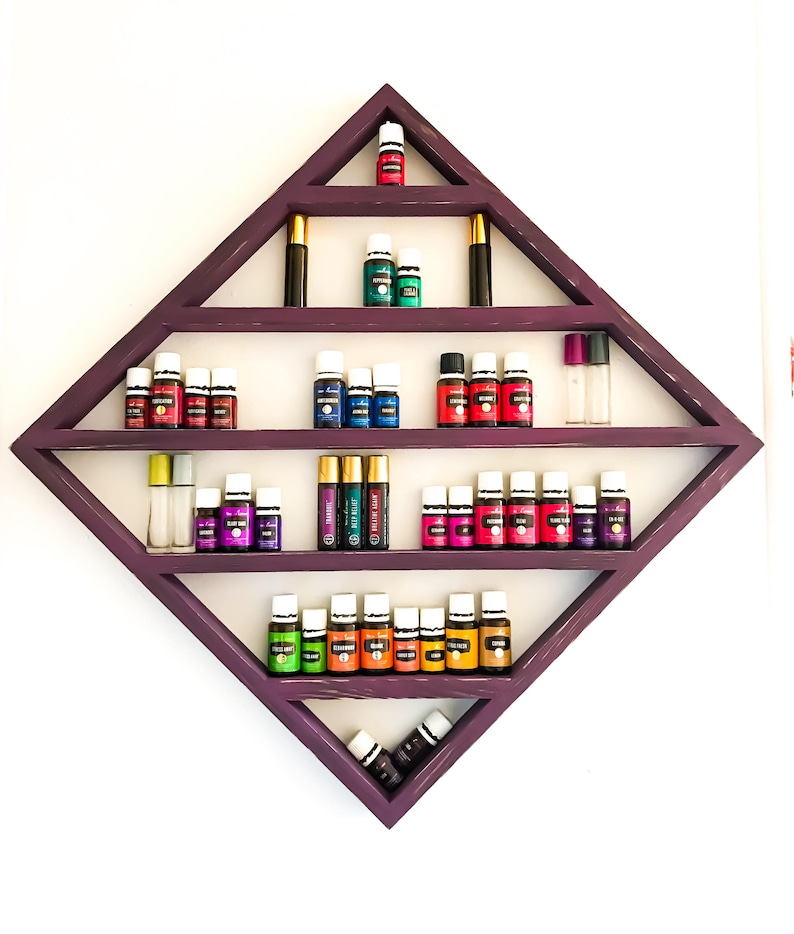 May include: A purple diamond-shaped shelf with five shelves holding various essential oil bottles. The bottles are in a variety of colours, including red, orange, yellow, green, blue, and purple. The shelf is mounted on a white wall.