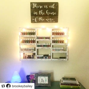 May include: A white wooden shelf with multiple rows of small glass bottles, each with a different colored liquid. The shelf is decorated with string lights and is mounted on a wall. A wooden sign above the shelf reads "There is oil in the house of the wise. Proverbs 21:20".