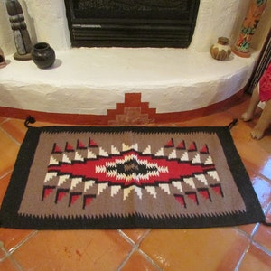 May include: A brown and black rug with a geometric pattern in red, white, and black. The rug has tassels on the ends and is laid out on a tiled floor.