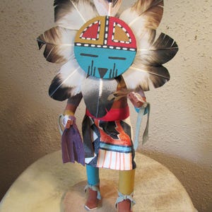 May include: A handcrafted kachina doll featuring a circular mask with turquoise, red, and yellow sections, surrounded by a large fan of black and white feathers. The figure wears a patterned skirt, has one blue and one yellow leg, and holds a purple cloth.